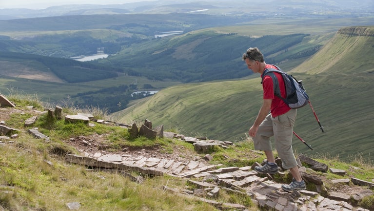A man walks the Brecon Beacons mountain range, the beautiful scenery pans out in the background as he walks upwards.